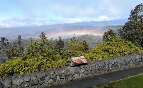 A scenic view of a volcanic landscape with a rainbow, framed by lush greenery and a stone wall.