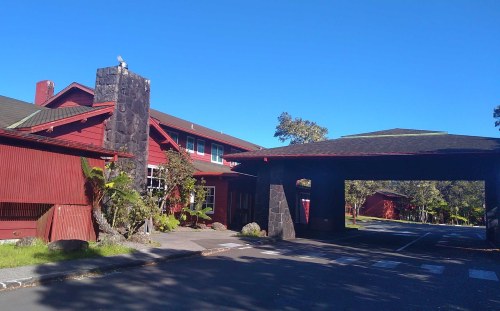 A red building with a stone chimney and a covered entrance, surrounded by greenery under a clear blue sky.