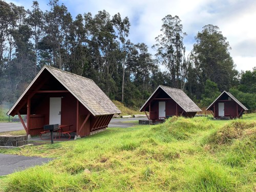 Three red cabins with sloped roofs in a grassy area, surrounded by trees under a cloudy sky.