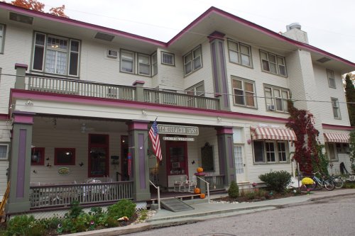 A charming inn with a welcoming porch, featuring pink accents and seasonal decorations, surrounded by greenery.