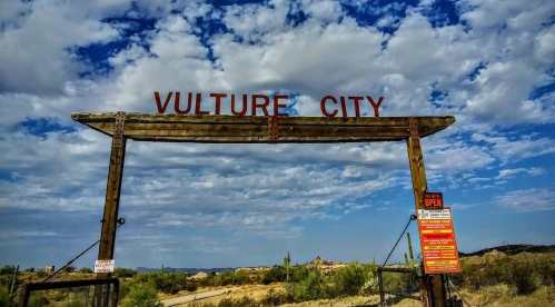 Wooden archway with "Vulture City" sign under a blue sky with clouds, surrounded by desert landscape.