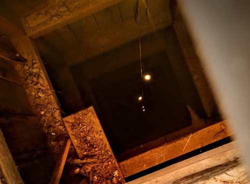 View down a dark well or shaft, with faint lights visible at the bottom and wooden beams surrounding the opening.