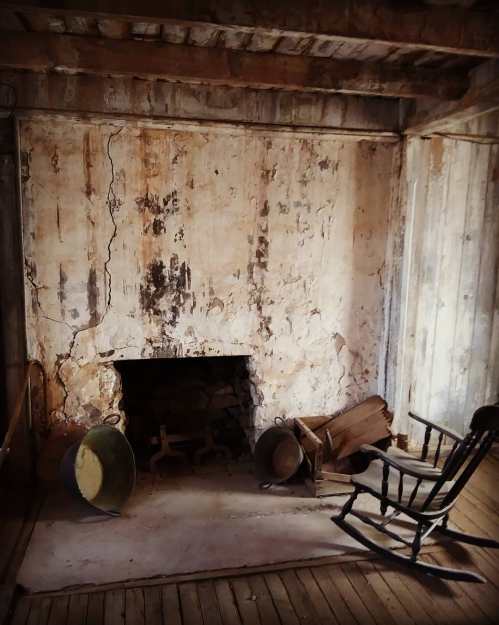 A rustic, empty room with cracked walls, a fireplace, and a wooden rocking chair.