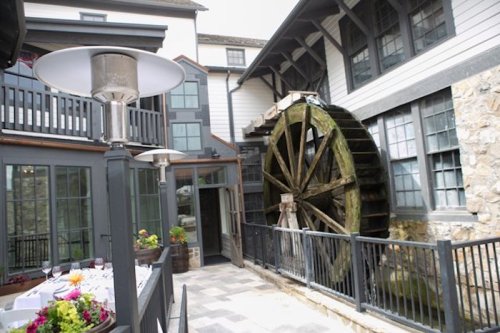 A rustic water wheel beside a modern building, with outdoor dining tables and greenery in a charming courtyard setting.