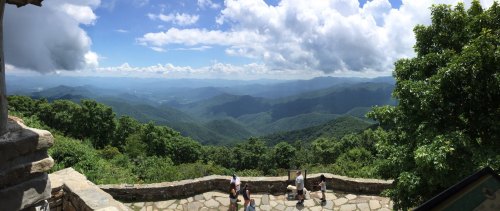 A panoramic view of lush green mountains under a blue sky with clouds, with people enjoying the scenery.