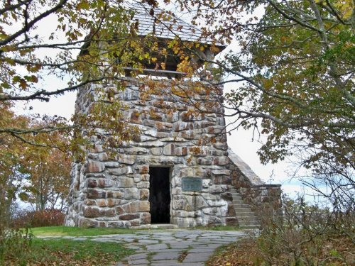 Stone tower surrounded by trees with autumn leaves, featuring a staircase and a plaque on the front.
