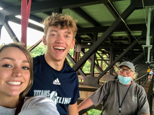 A young man and woman smile for a selfie with an older woman in a mask, all on a bridge with greenery in the background.