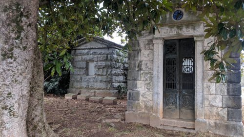 Two stone mausoleums under a large tree, one with ornate doors and the other simpler, surrounded by grass and foliage.