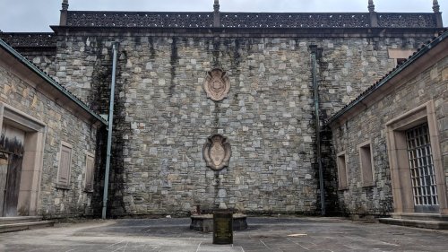 Stone wall with two decorative plaques, surrounded by buildings and a paved area, under a cloudy sky.