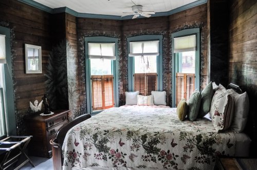 Cozy bedroom with a floral quilt, wooden walls, and large windows, featuring a bedside table and decorative pillows.