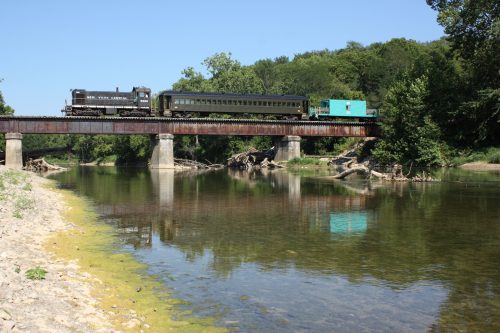 A train crosses a bridge over a calm river, surrounded by lush greenery and clear blue skies.