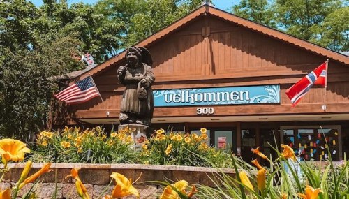 A wooden building with "Velkommen" sign, featuring a statue and flowers in front, with American and Norwegian flags.
