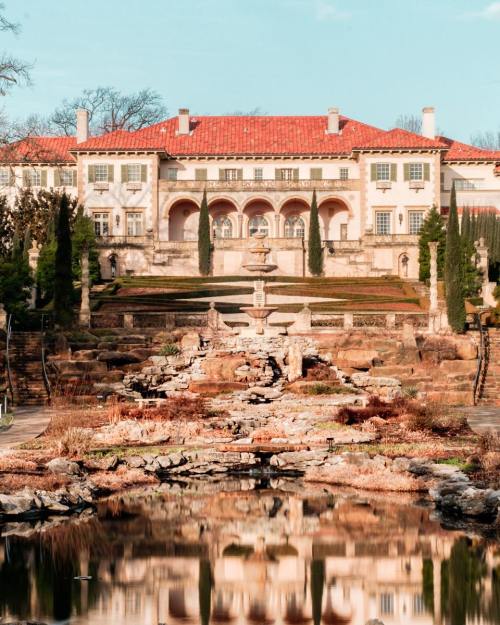 A grand mansion with a red-tiled roof, surrounded by gardens and a reflecting pond, under a clear blue sky.