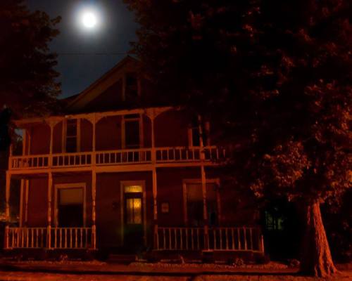 A dimly lit house at night, illuminated by a full moon, with trees casting shadows in the foreground.
