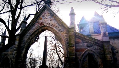 Stone archway of a cemetery with the word "Cemetery" engraved, surrounded by bare trees and a bright sky.