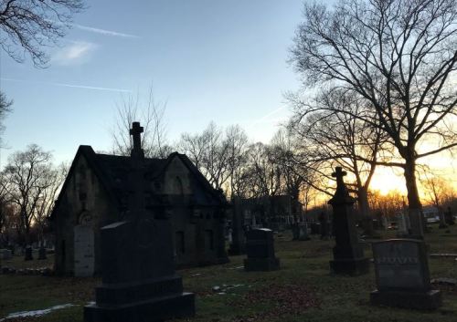 A cemetery at sunset, featuring gravestones and a mausoleum surrounded by bare trees.