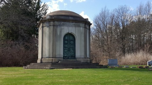 A stone mausoleum with a domed roof and ornate door, surrounded by trees and grass in a cemetery setting.
