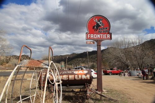 A roadside sign for "Frontier" with a cowboy graphic, next to an old gas tank and a museum, set against a cloudy sky.