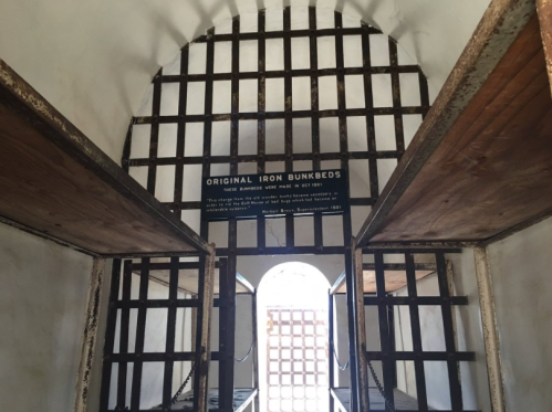 Interior view of a historic prison cell with iron bunk beds and barred windows, featuring a sign about their origin.