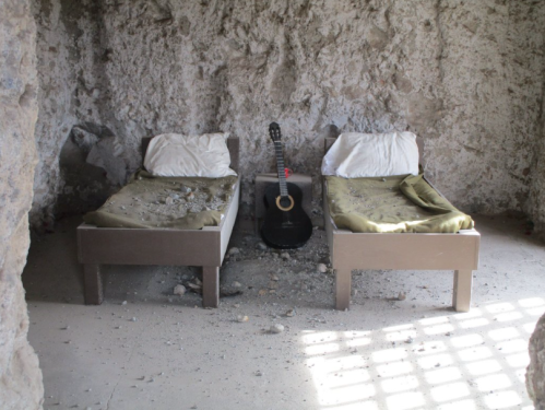 Two simple beds with unmade sheets in a stone room, with a guitar leaning against the wall between them.