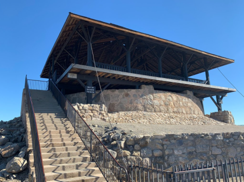 A stone building with a wooden roof, situated on a rocky hill, with stairs leading up to it under a clear blue sky.