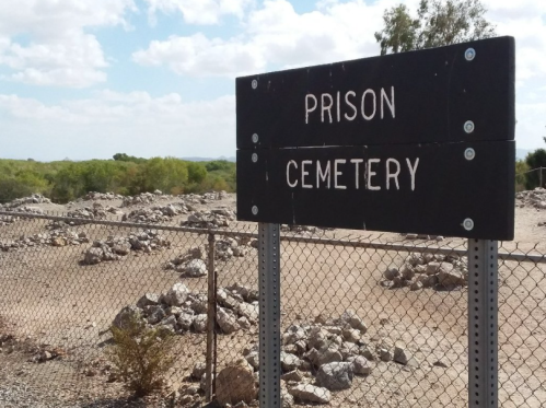Sign reading "Prison Cemetery" next to a rocky, fenced area with sparse vegetation under a cloudy sky.