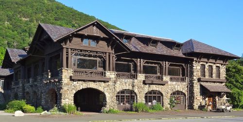 A large, rustic building with stone and wooden architecture, surrounded by greenery and mountains in the background.