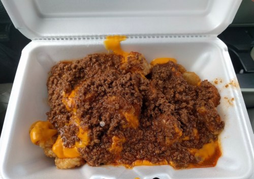 A takeout container filled with tater tots topped with chili and melted cheese.