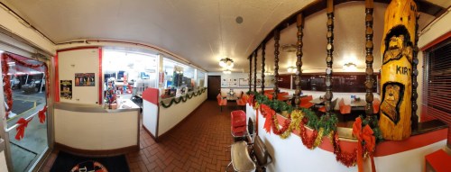 A cozy diner interior decorated with festive garlands and lights, featuring a wooden statue and red seating.