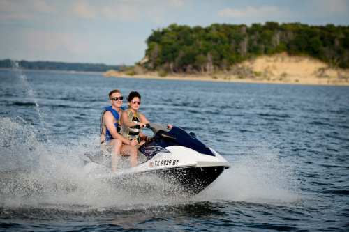 Two people ride a jet ski on a lake, splashing water, with a wooded shoreline in the background.