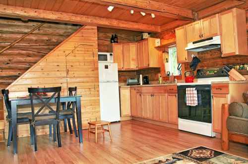 Cozy log cabin kitchen with wooden cabinets, a dining table, and a staircase, featuring modern appliances and warm lighting.