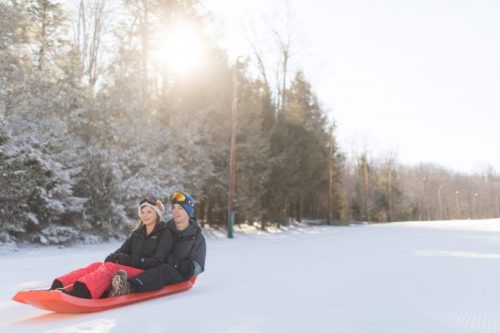 A couple sits on a sled in a snowy landscape, surrounded by trees, enjoying a sunny winter day.