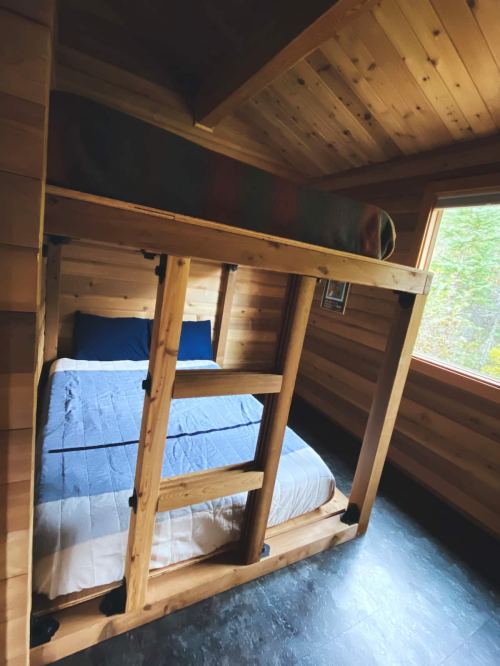 Cozy wooden bedroom featuring a lofted bed with blue bedding and a large window overlooking greenery.