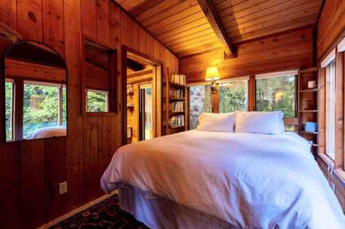 Cozy wooden bedroom with a large bed, bookshelves, and windows overlooking greenery. Warm, inviting atmosphere.