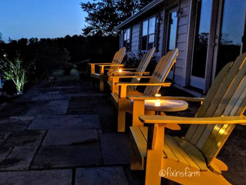 Four yellow Adirondack chairs on a stone patio, illuminated by candlelight, with a twilight sky in the background.