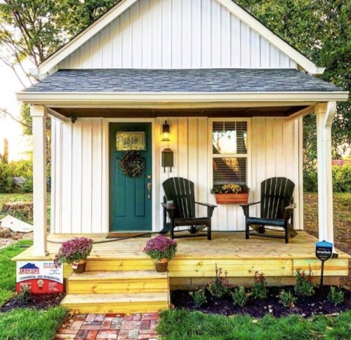 A charming white house with a blue door, two black chairs on the porch, and colorful flowers in pots.
