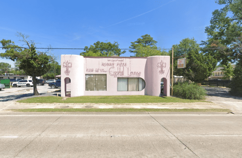 A pink building with a curved design, featuring a sign for Roman Pizza and surrounded by trees and greenery.