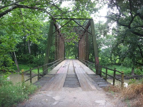 A weathered metal bridge surrounded by lush greenery, leading over a calm waterway.