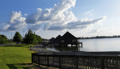 A serene lakeside view with wooden structures, green grass, and fluffy clouds in a blue sky.