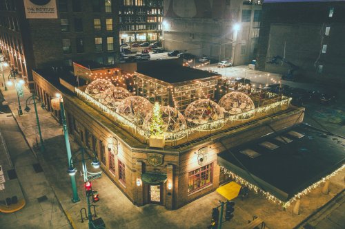 Aerial view of a festive restaurant with illuminated domes and string lights on the rooftop, surrounded by city buildings.