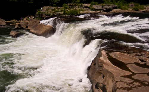 A cascading waterfall flows over rocky ledges, surrounded by lush greenery and a serene river landscape.