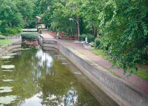 A serene canal scene with a path, greenery, and a couple walking near the water's edge.