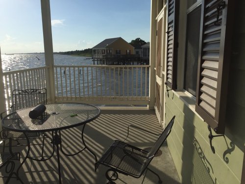 A balcony with a table and chairs overlooking a calm waterfront and colorful houses on stilts in the distance.