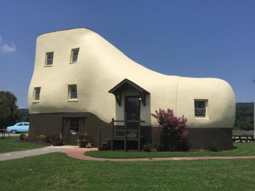 A whimsical shoe-shaped house with a brown base and light-colored upper, surrounded by greenery and flowers.