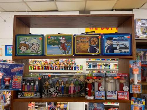 Display of colorful PEZ lunchboxes and various PEZ dispensers on a wooden shelf in a store.