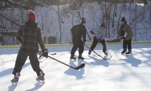 A group of children playing ice hockey on a snowy outdoor rink, wearing winter clothing and skating gear.