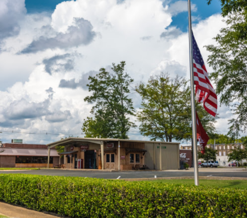 A small building with a wooden facade, surrounded by trees and a flagpole, under a partly cloudy sky.
