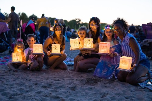 A group of women and children holding glowing lanterns on a beach at dusk, smiling and enjoying the evening.