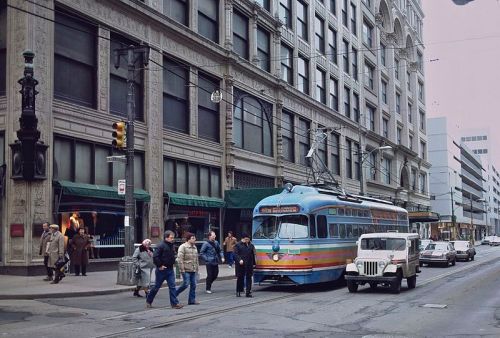 A colorful streetcar stops on a city street, with pedestrians and vintage cars nearby, set against historic buildings.