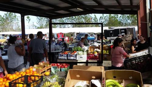 A bustling market scene with people shopping for fresh produce and various goods under a large canopy.